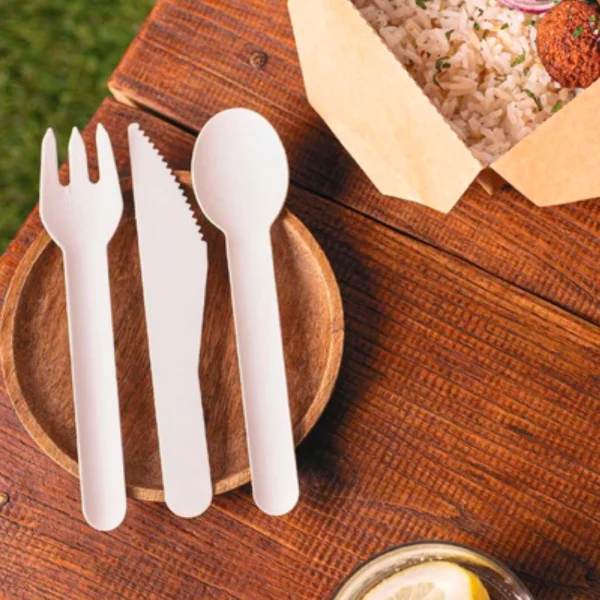 Cubiertos descartables de papel ecológicos (tenedor, cuchillo, cuchara) sobre plato de madera, junto a envase para llevar.