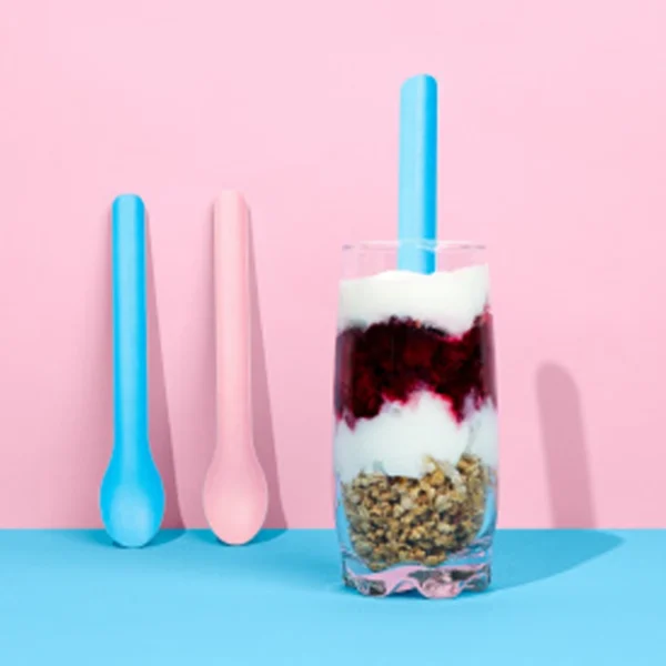 Cucharas largas descartables de papel en tonos pastel. Cuchara azul y cuchara rosa en vaso con yogur y granola. Alternativa sostenible.