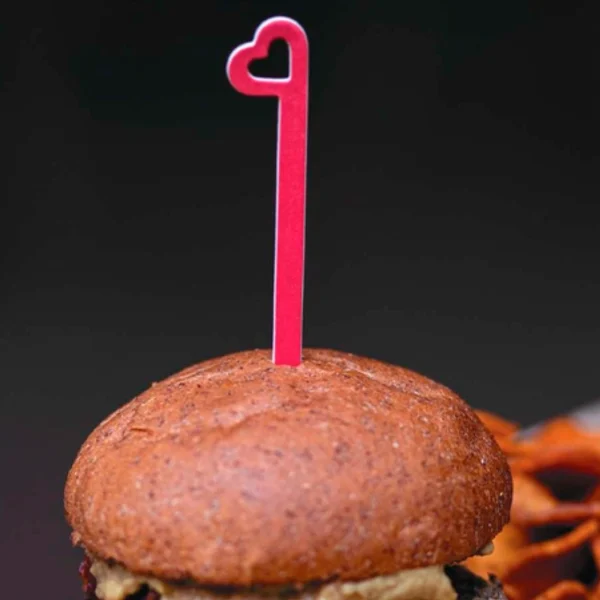 Palillo decorativo de papel rojo con forma de corazón en una hamburguesa. Ideal para eventos sostenibles y servicios de comida ecológicos.